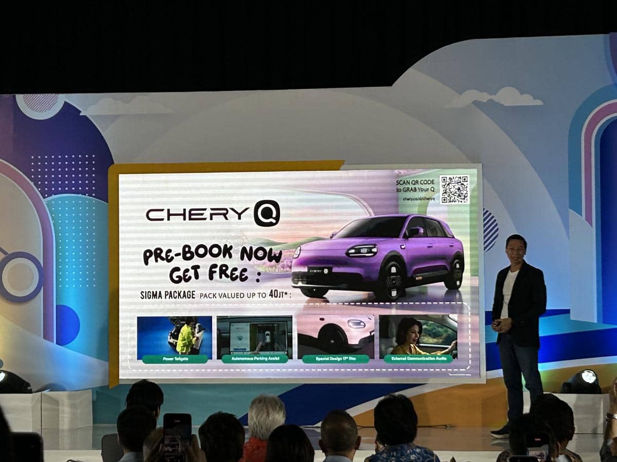 Seorang pembicara berdiri di samping layar besar yang menampilkan promosi pre-book mobil Chery Q berwarna ungu dengan berbagai penawaran bonus.