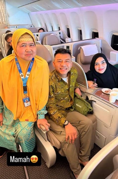 Seorang pria dan wanita duduk di kursi pesawat kelas bisnis bersama seorang perempuan berkerudung kuning yang berdiri di samping mereka.