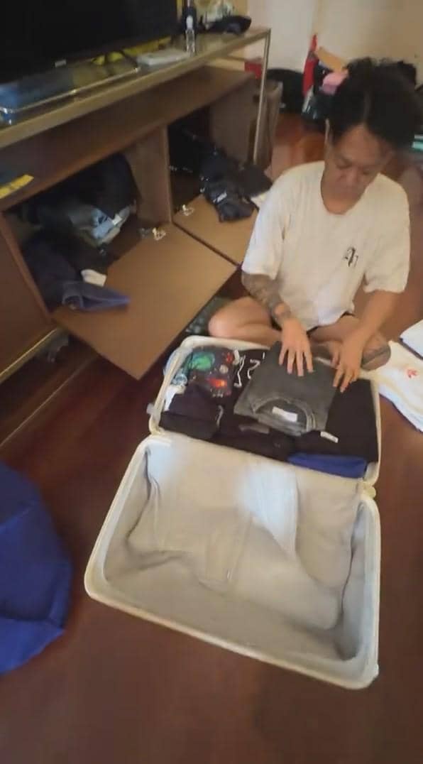 Seorang pria duduk di lantai sedang melipat dan menata pakaian ke dalam koper besar di kamar dengan lantai kayu.