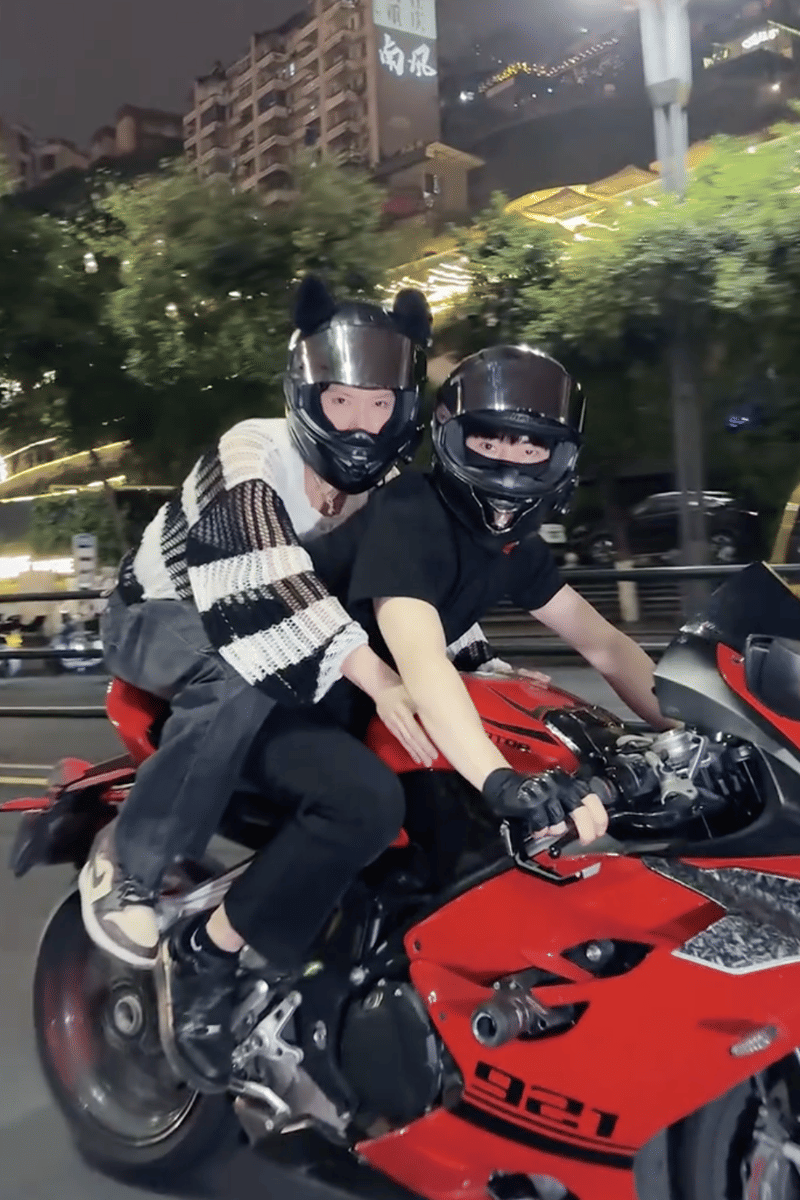Dua orang mengenakan helm hitam sedang mengendarai motor sport merah di jalan kota pada malam hari dengan latar gedung bertingkat.