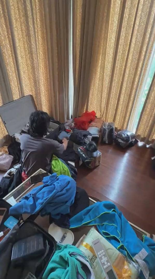 Seseorang duduk di lantai kamar dengan koper terbuka di sekeliling pakaian dan barang-barang yang berserakan di lantai kayu.