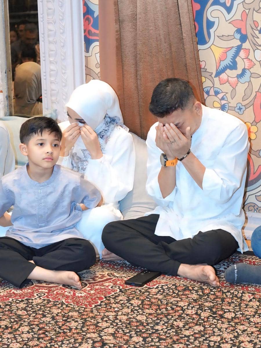 Sepasang suami istri berpakaian putih duduk di atas karpet bersama anak laki-laki, tampak berdoa sebelum keberangkatan haji.