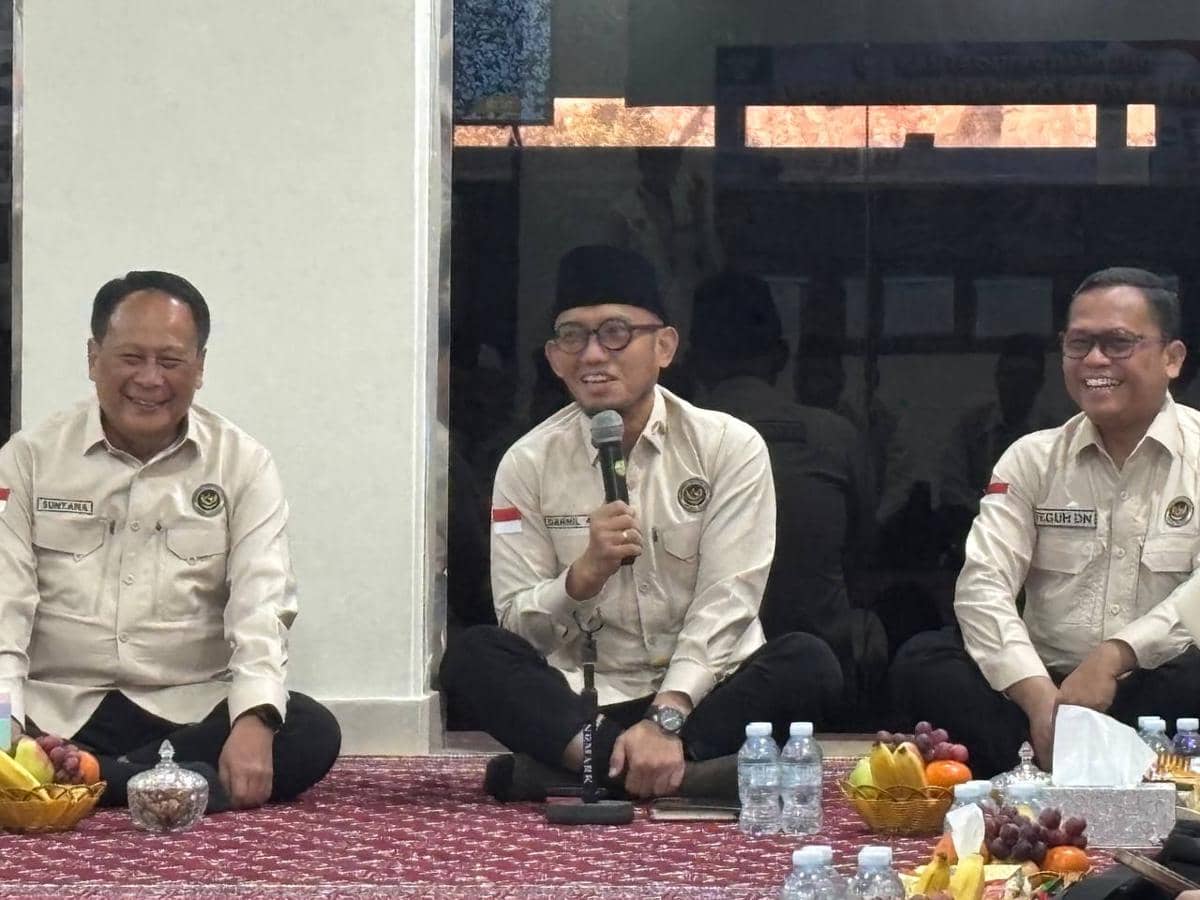 Tiga pria berpakaian seragam krem duduk bersila di atas karpet dengan hidangan buah dan air mineral di depan mereka, salah satunya memegang mikrofon.