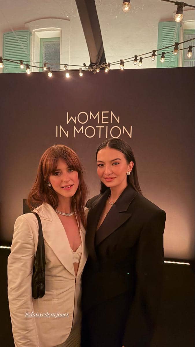 Dua perempuan berpose bersama di acara Women in Motion dengan latar bertuliskan nama acara dan pencahayaan hangat di sekitarnya.