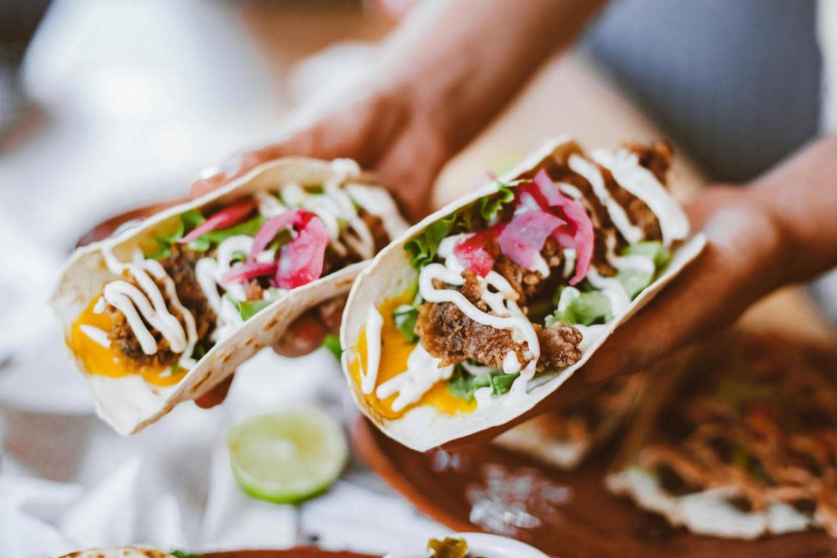 Dua taco berisi daging, sayuran segar, saus keju, dan krim putih dipegang dengan tangan di atas meja makan.