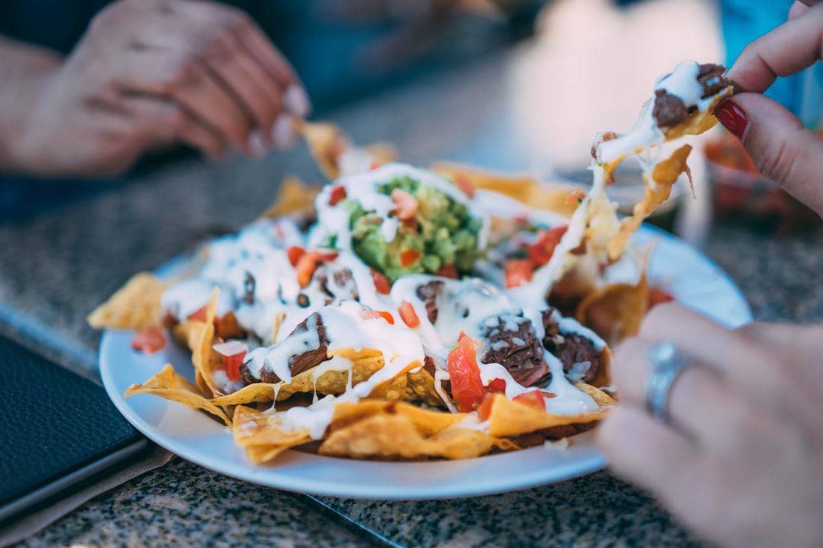 Piring berisi nachos dengan topping daging, saus keju, krim putih, potongan tomat, dan guacamole di atas meja makan.