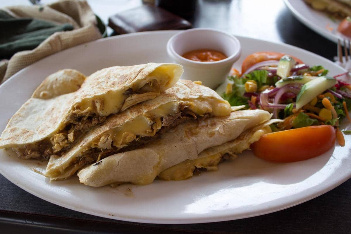 Quesadilla berisi ayam dan keju leleh disajikan di atas piring putih dengan saus celup dan salad segar berisi sayuran serta tomat.
