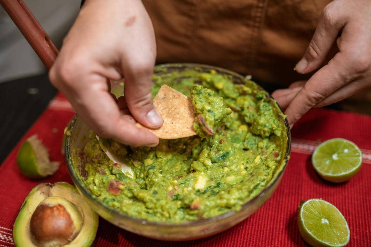 Seseorang mencelupkan keripik tortilla ke dalam mangkuk berisi guacamole segar dengan potongan alpukat dan jeruk nipis di sekitarnya.