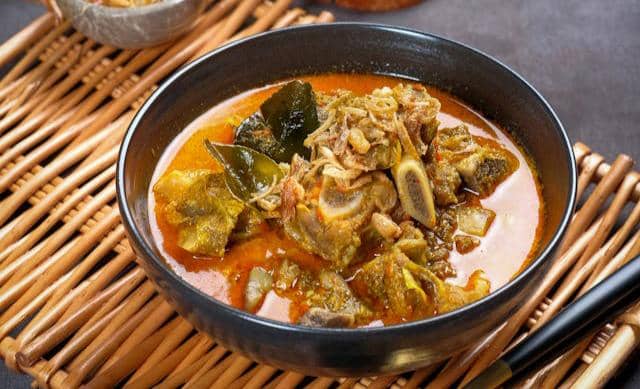 Semangkuk gulai kambing khas dengan kuah santan berwarna kuning kemerahan, potongan daging dan tulang kambing, serta taburan bawang goreng.