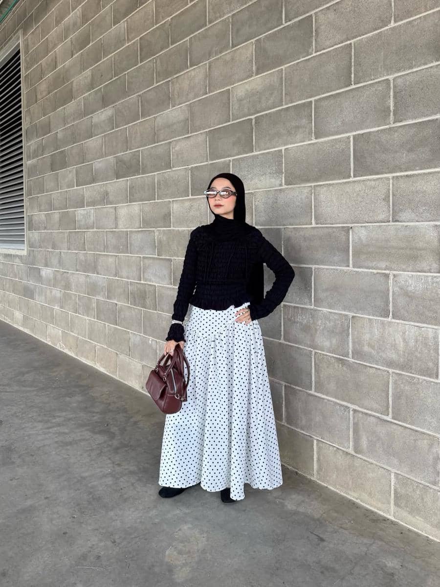 Seorang wanita berhijab mengenakan atasan hitam dan rok panjang putih bermotif polkadot sambil memegang tas cokelat di depan dinding bata abu-abu.