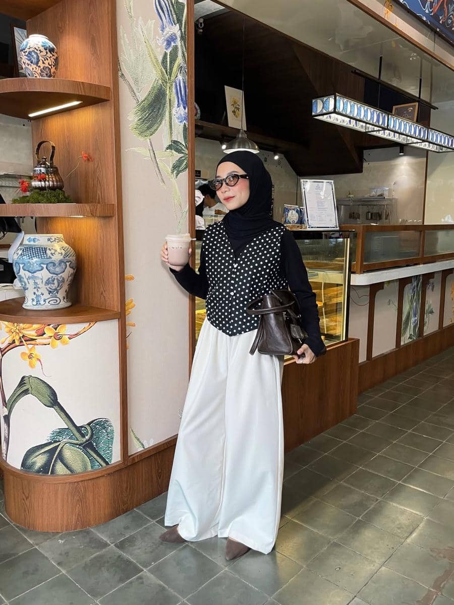 Seorang wanita berhijab mengenakan vest hitam bermotif polkadot dan celana putih lebar sambil memegang minuman di kafe bergaya modern.