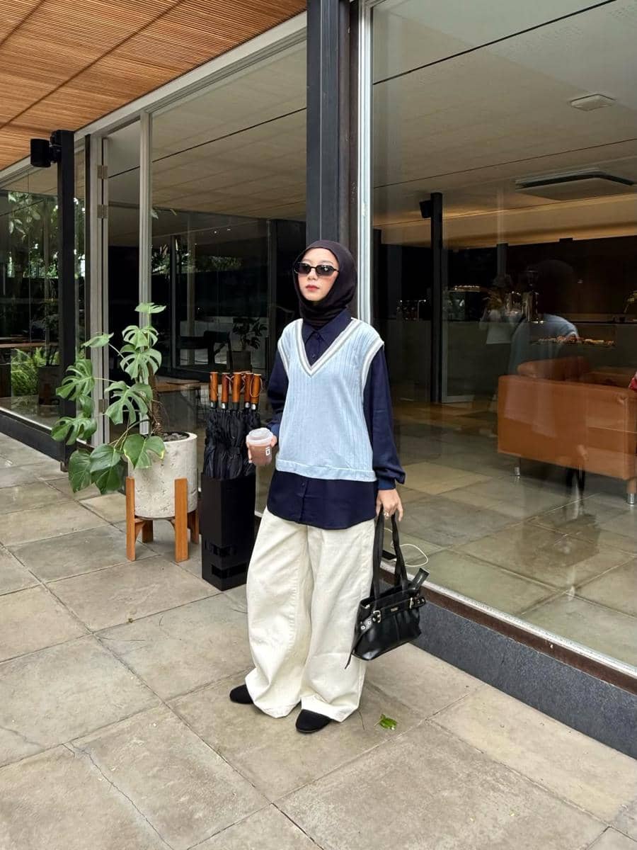 Seorang wanita berhijab mengenakan vest rajut biru muda di atas kemeja hitam, celana panjang krem, dan membawa tas hitam di area luar kafe.