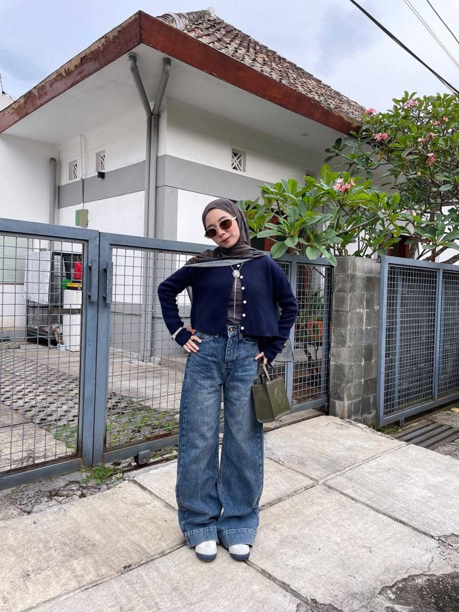 Seorang wanita berhijab bergaya kasual mengenakan cardigan biru tua dan celana jeans longgar, berpose di depan pagar rumah dengan tanaman hijau di sampingnya.