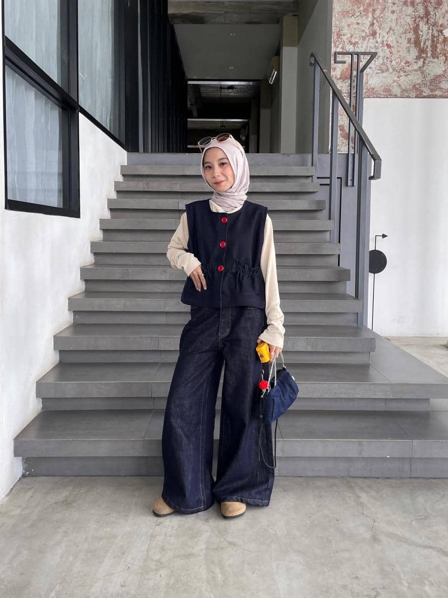 Seorang wanita berhijab mengenakan vest biru tua dengan kancing merah, celana jeans lebar, dan tas kecil di tangga beton modern.