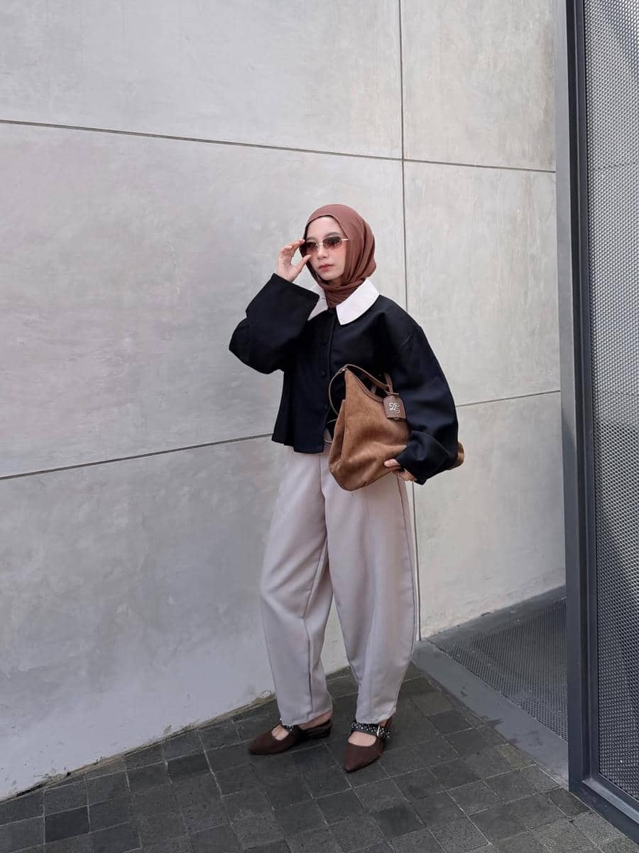Seorang wanita berhijab bergaya kasual mengenakan atasan hitam berkerah putih, celana longgar krem, dan tas cokelat di area outdoor.