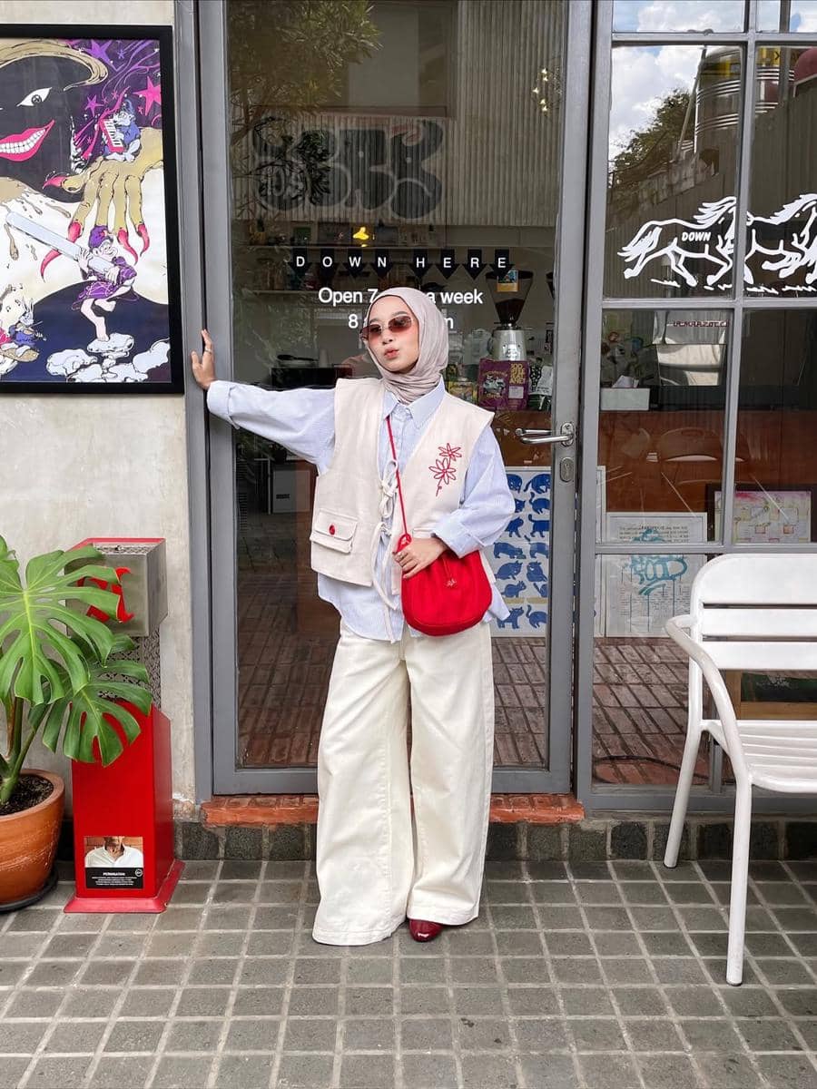 Seorang wanita berhijab mengenakan vest krem, kemeja biru muda, celana panjang putih lebar, dan tas selempang merah di depan kafe.