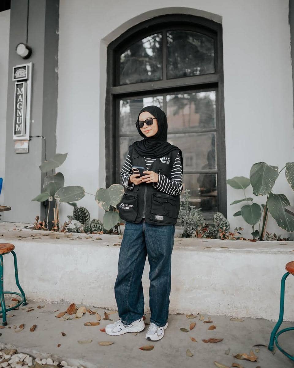 Seorang wanita berhijab mengenakan vest hitam, kaus garis-garis, celana jeans longgar, dan sepatu putih berdiri di depan jendela besar.