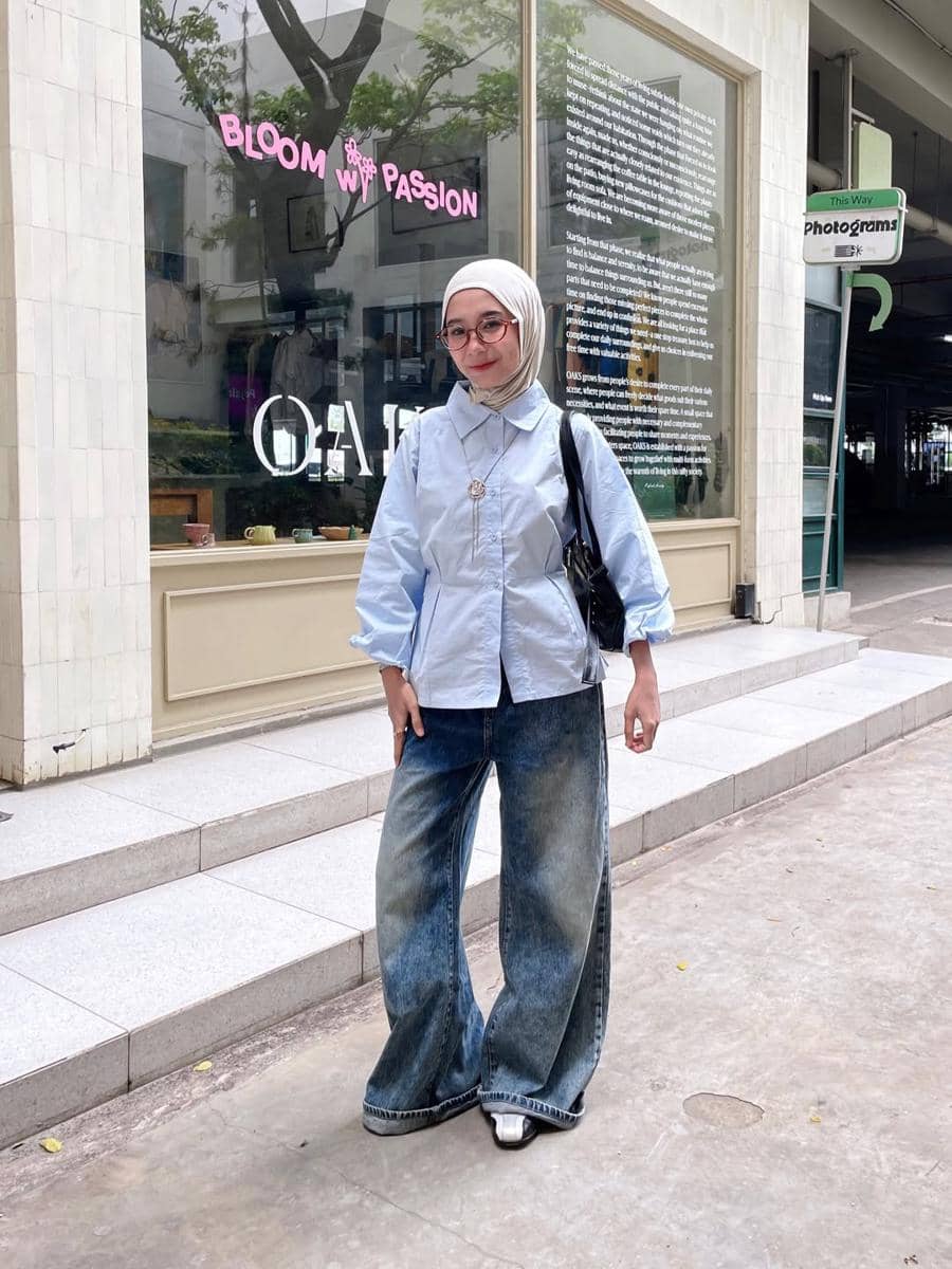 Seorang wanita berhijab bergaya kasual mengenakan kemeja biru muda dan celana jeans wide leg di depan toko dengan jendela besar.