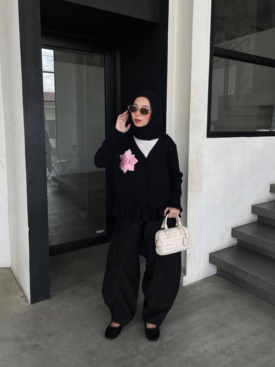 Seorang wanita berhijab bergaya kasual mengenakan kardigan hitam dengan hiasan bunga pink, celana longgar hitam, dan tas kecil krem.