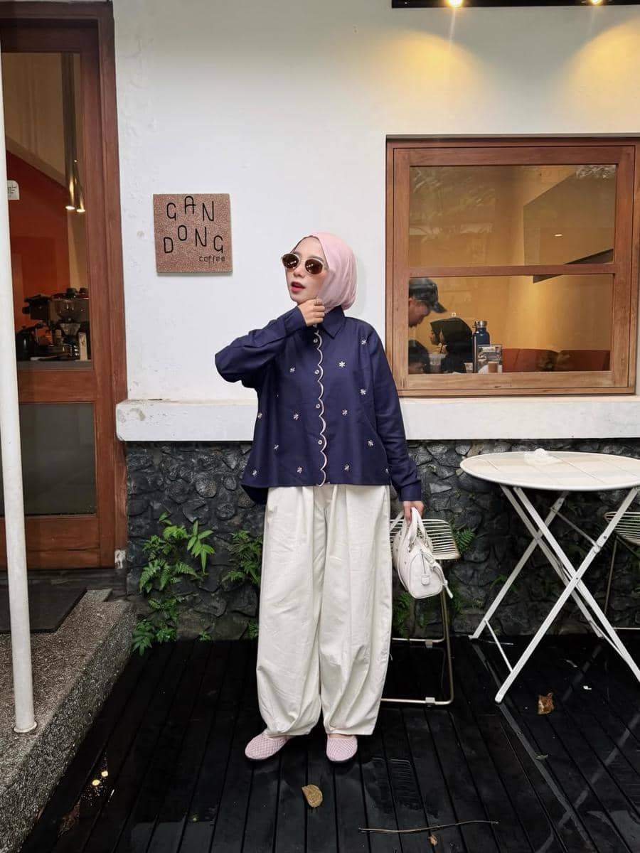 Seorang wanita berhijab mengenakan kemeja biru tua dan celana putih longgar berpose di depan kafe dengan gaya kasual modern.