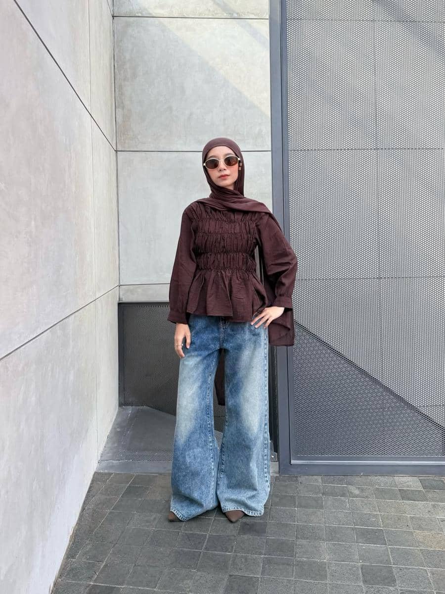 Seorang wanita berhijab mengenakan atasan cokelat dengan detail kerut dan celana jeans longgar, berpose di depan dinding abu-abu modern.