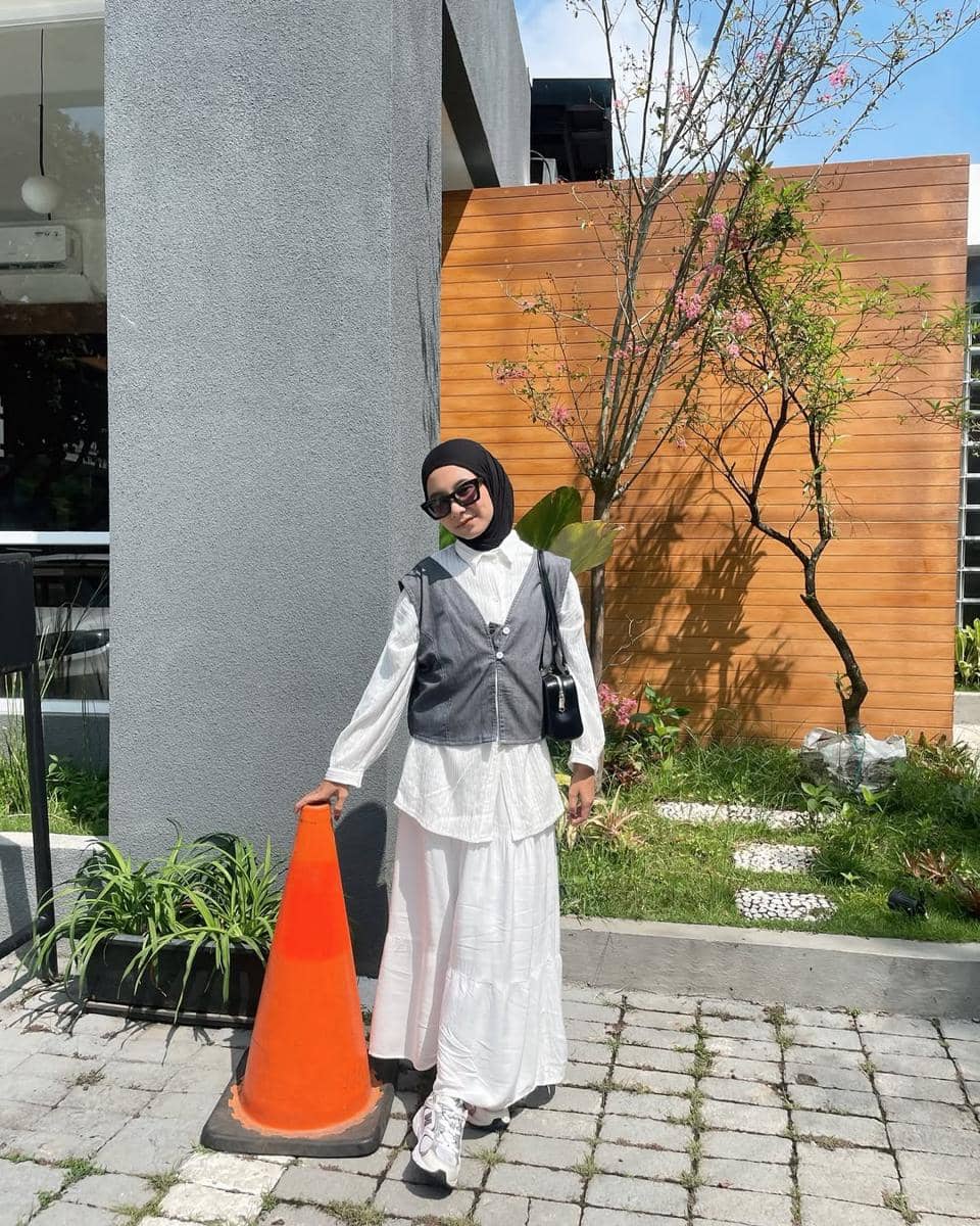 Seorang wanita berhijab mengenakan vest abu-abu di atas kemeja putih panjang dan rok putih, berdiri di samping cone oranye di area outdoor.