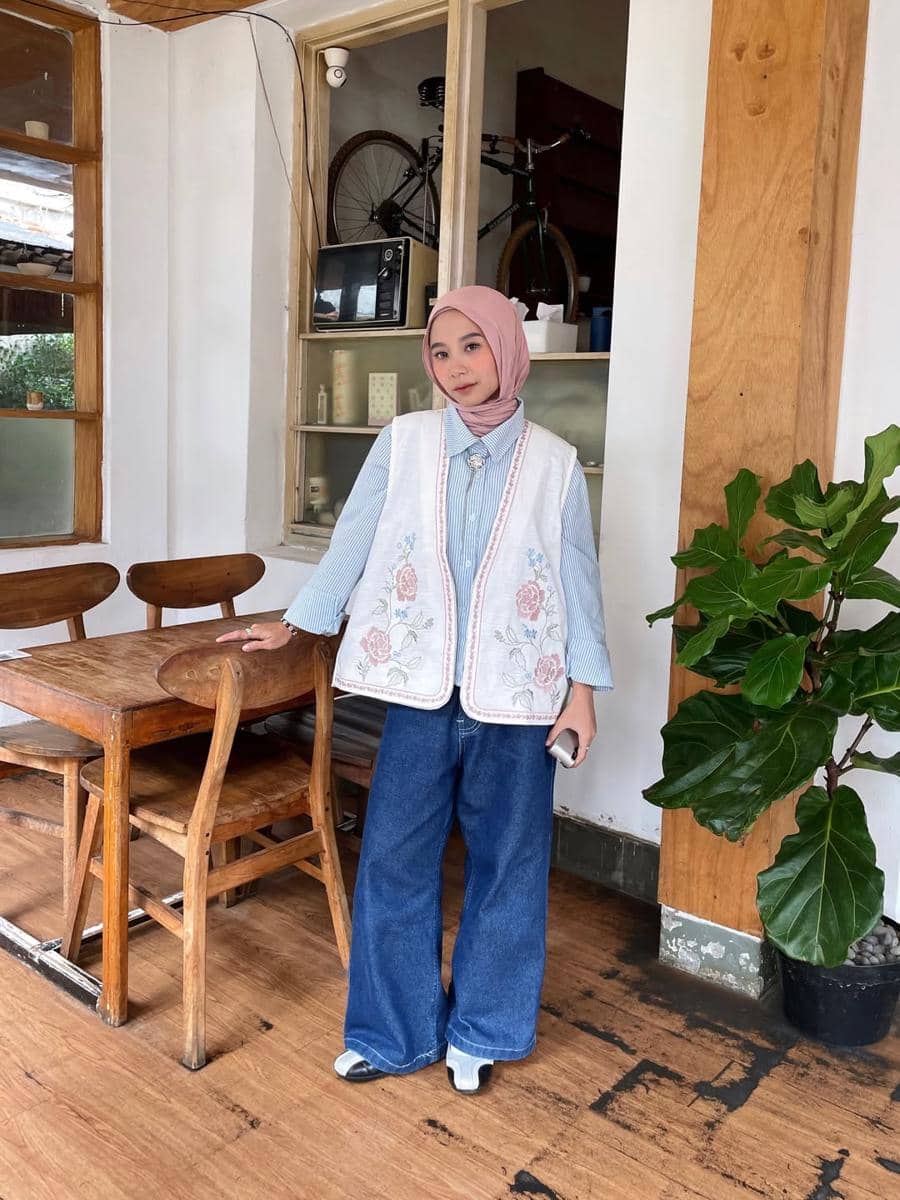 Seorang wanita berhijab mengenakan vest bermotif bunga, kemeja biru bergaris, dan celana jeans lebar berdiri di dalam kafe bergaya rustic.