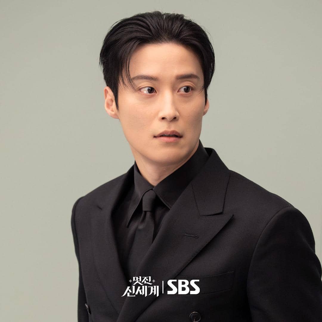 Seorang pria mengenakan setelan jas hitam dan dasi hitam dengan latar belakang abu-abu muda dalam still cut drama My Royal Nemesis.