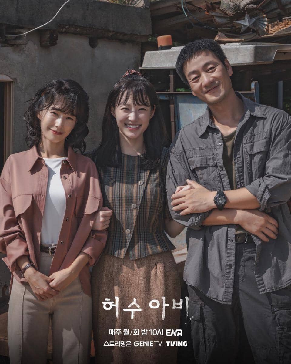 Tiga pemeran utama drama Korea The Scarecrow berdiri bersama sambil tersenyum di luar ruangan dengan latar bangunan sederhana.