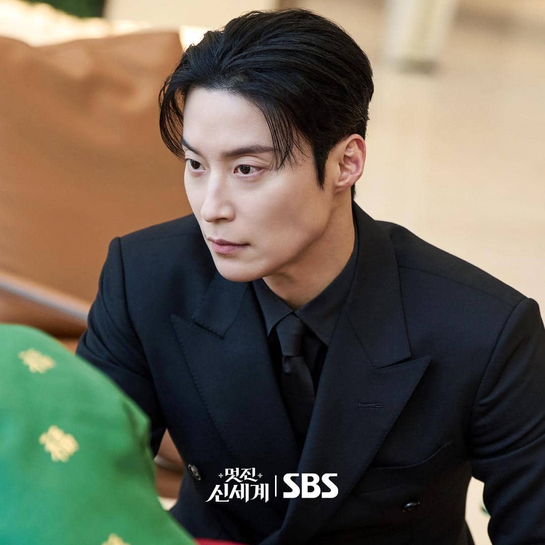 Seorang pria berpakaian jas hitam duduk dengan ekspresi serius dalam adegan drama My Royal Nemesis produksi SBS.