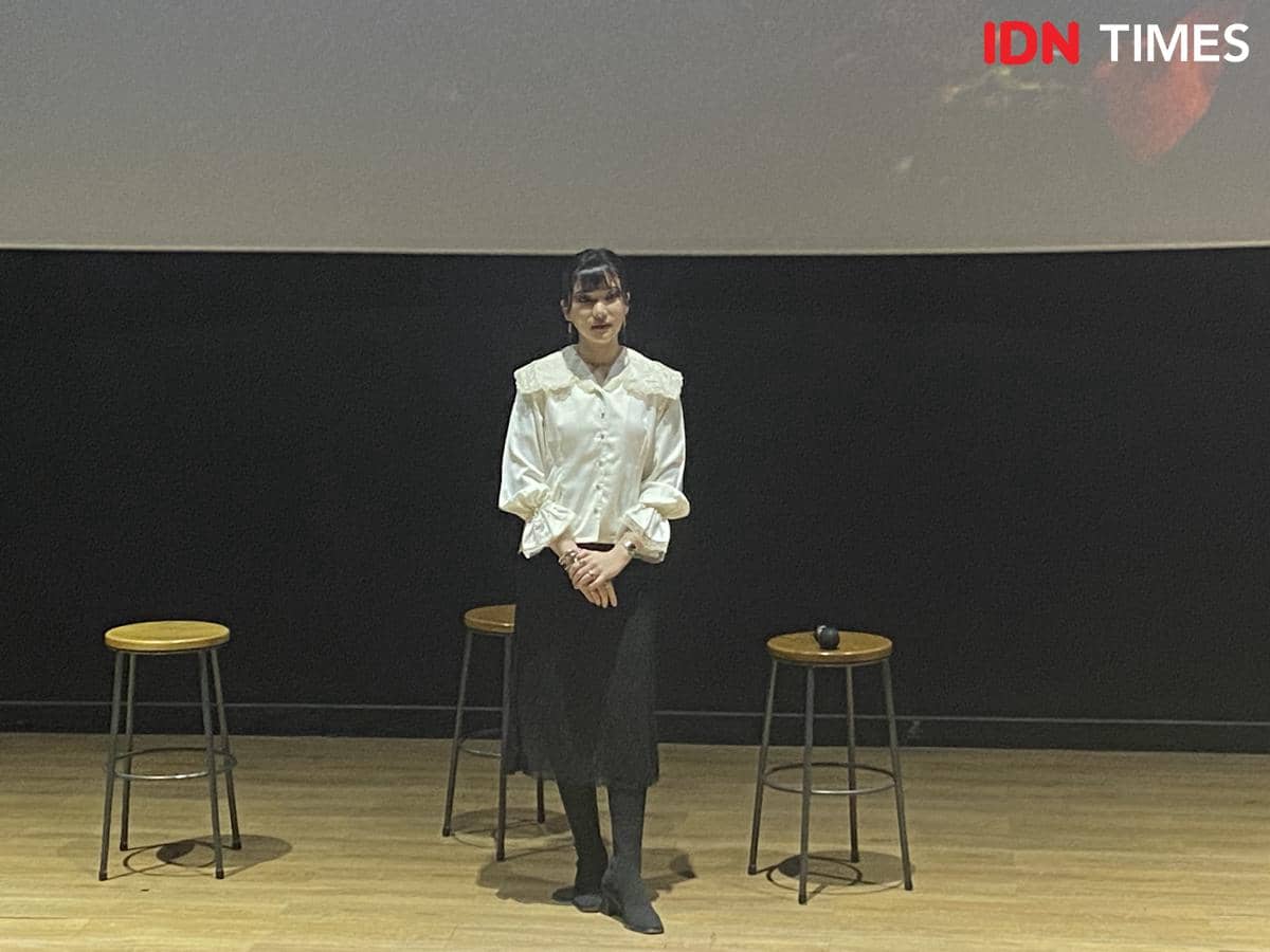 Seorang perempuan berdiri di atas panggung kayu dengan latar layar besar dan dua kursi tinggi di acara Media Day album EKLEKTIKO di CGV fX Sudirman.
