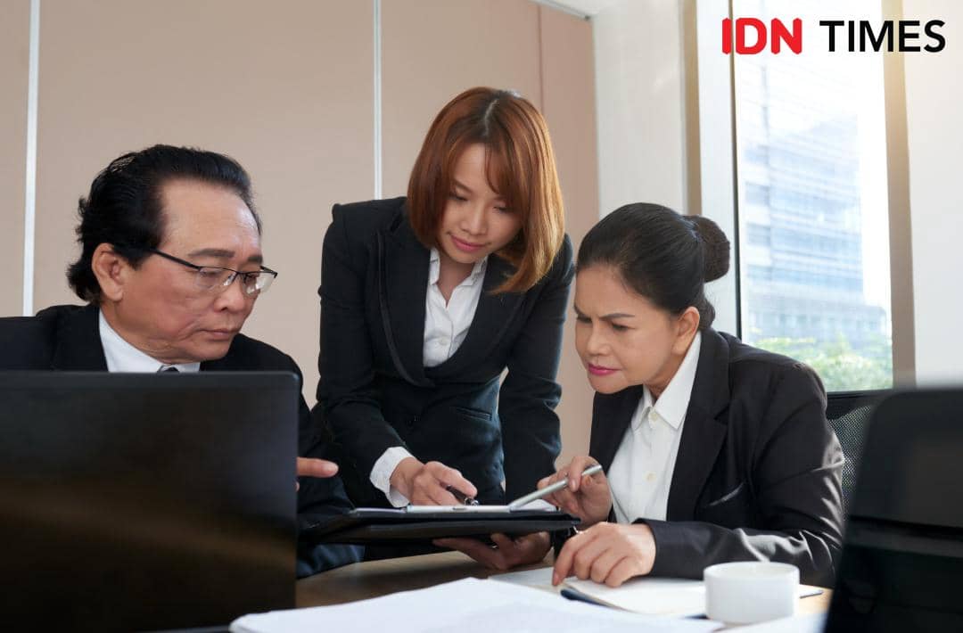 Tiga profesional hukum berdiskusi di ruang kantor modern sambil meninjau dokumen dan menggunakan laptop di meja kerja.