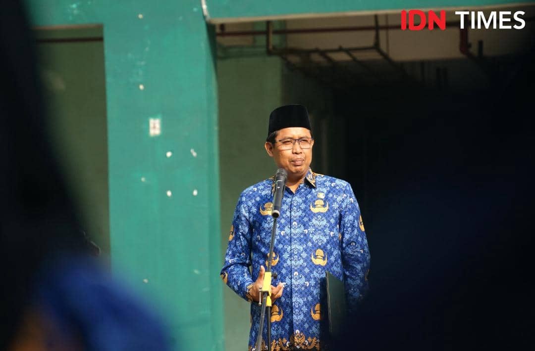 Prof. Ridwan berbicara di depan mikrofon di sebuah ruangan dengan latar hijau.