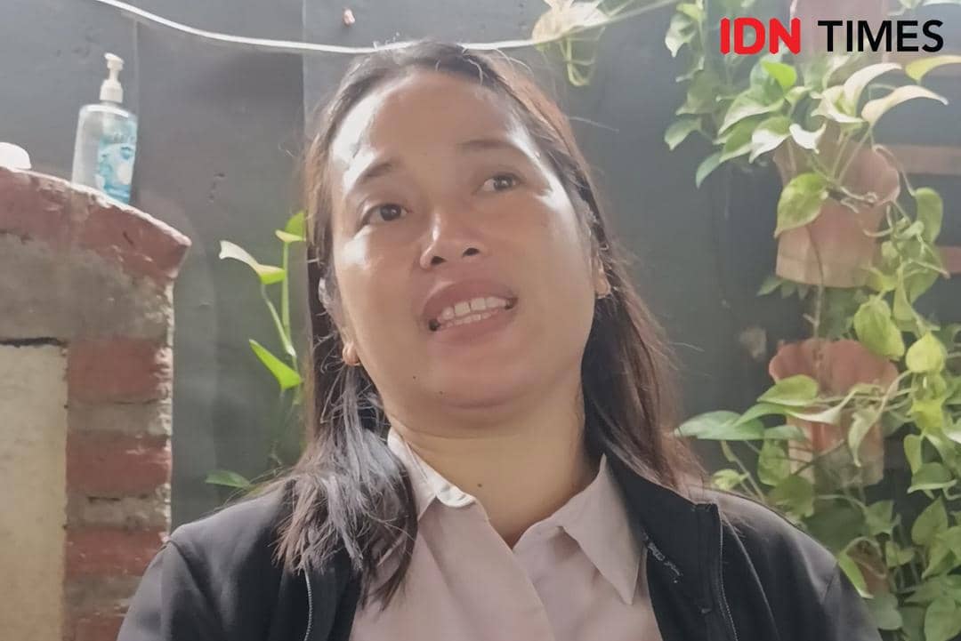 Seorang perempuan berbicara dengan ekspresi serius di area luar ruangan dengan latar tanaman hijau dan dinding bata.