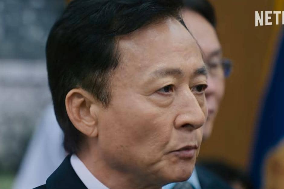 Seorang aktor pria dalam adegan drama Korea Teach You a Lesson sedang berbicara dengan ekspresi serius di depan umum.