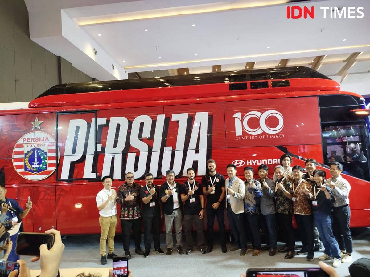 Bus listrik merah bertuliskan Persija Jaya Raya diperkenalkan bersama sekelompok orang yang berpose di depannya di sebuah acara peluncuran.