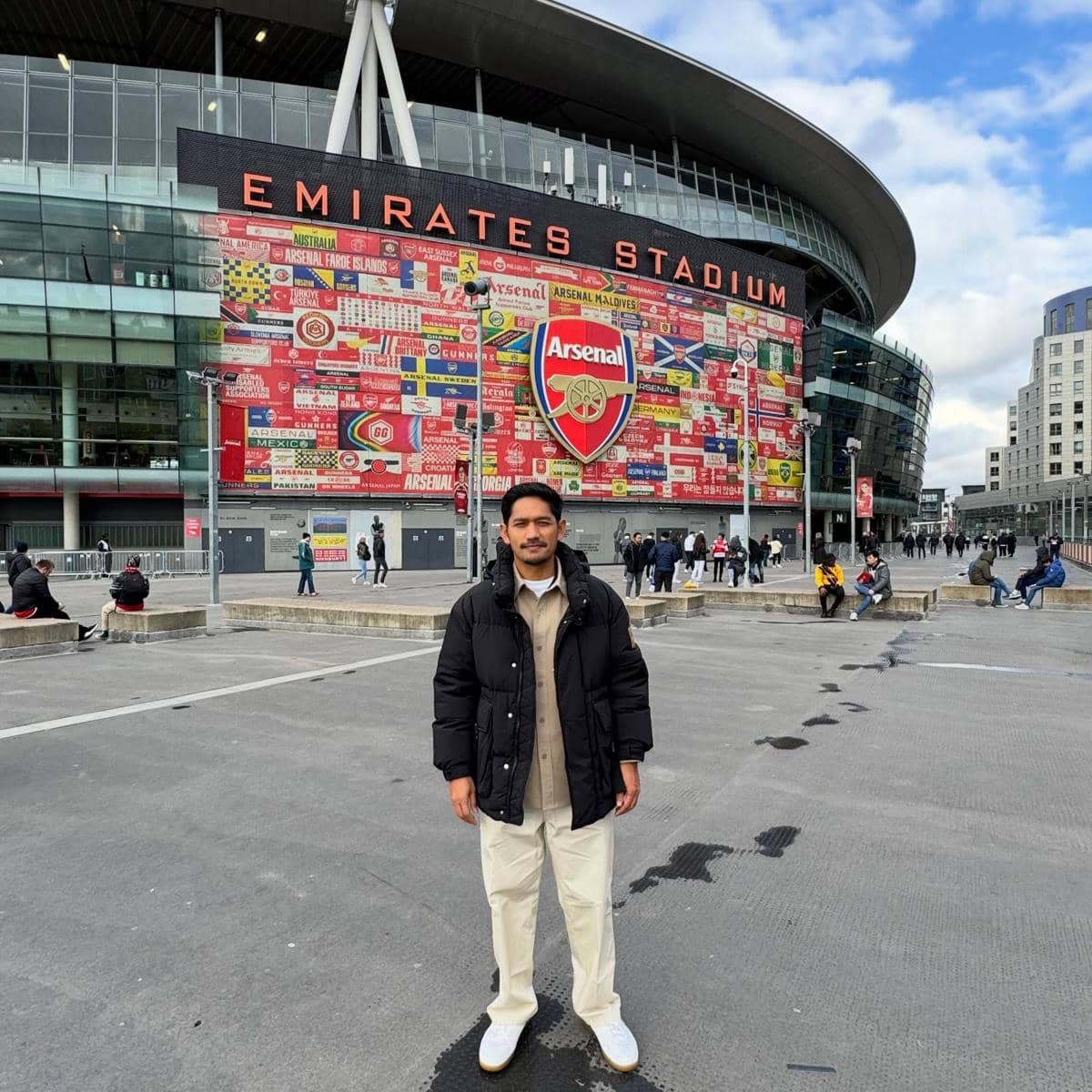 Seorang pria berdiri di depan Emirates Stadium dengan latar belakang logo besar Arsenal dan dinding bergambar bendera warna merah.