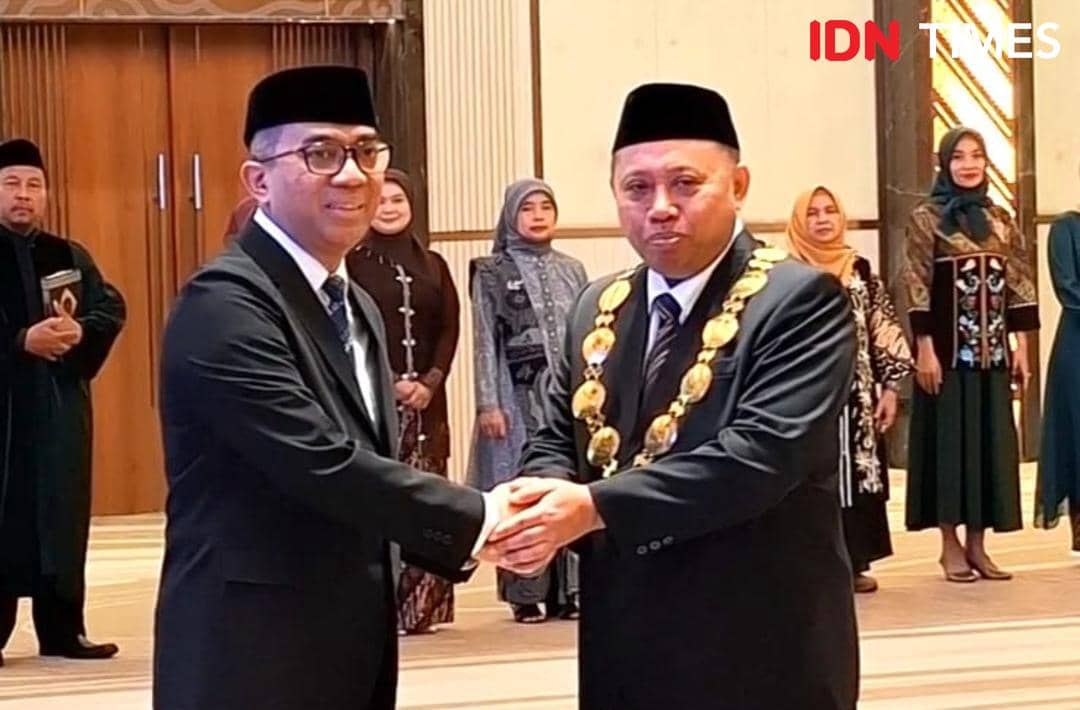 Dua pria mengenakan setelan jas dan peci berjabat tangan di acara resmi, salah satunya memakai kalung jabatan emas, disaksikan beberapa orang di belakang.