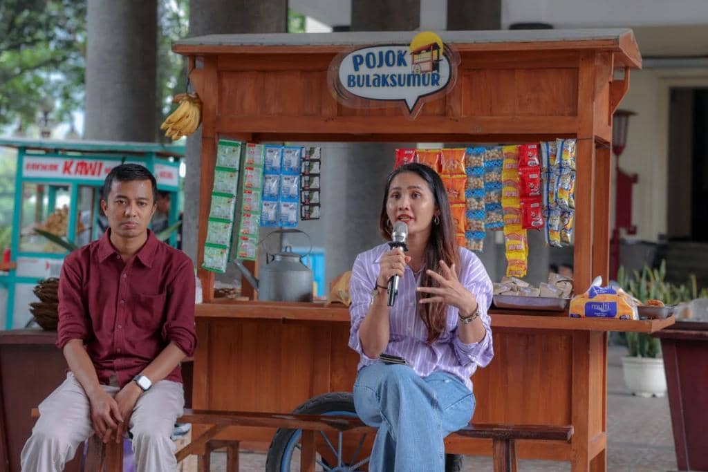 Seorang perempuan berbicara dengan mikrofon di depan warung kayu bertuliskan Pojok Bulaksumur, duduk di samping seorang pria berbusana kasual.