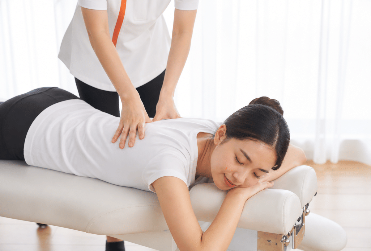 Seorang terapis melakukan pijat punggung pada perempuan yang berbaring santai di atas meja terapi dalam suasana ruangan terang.