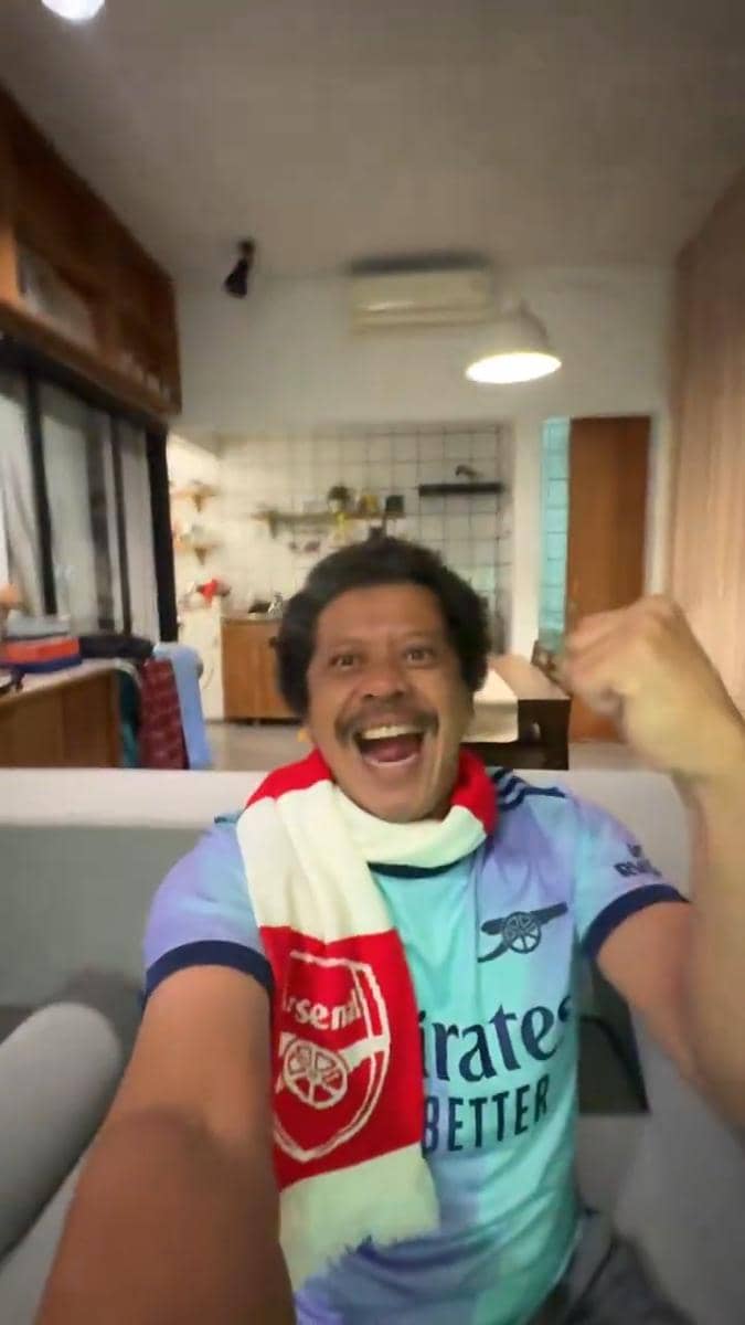 Seorang pria mengenakan jersey dan syal Arsenal tersenyum lebar sambil mengepalkan tangan di ruang tamu yang terang.