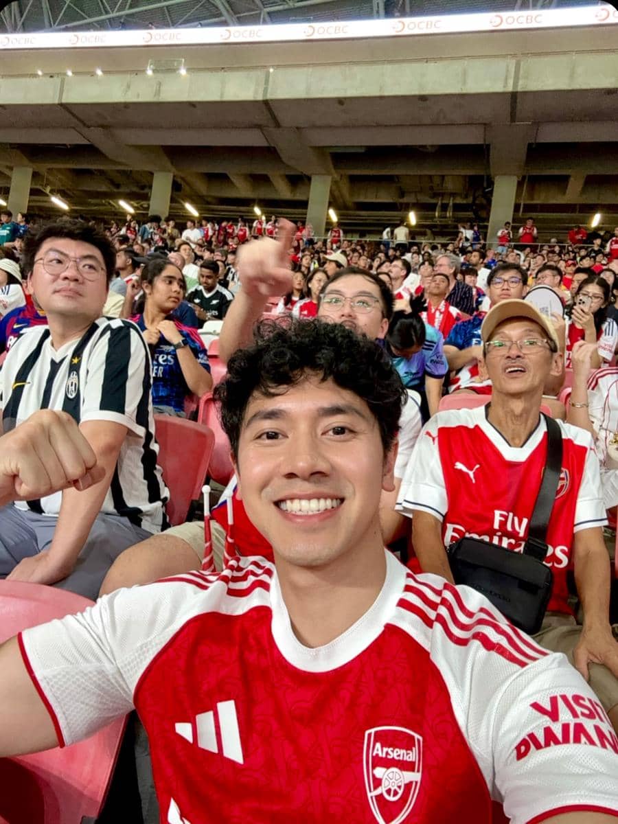 Seorang pria mengenakan jersey Arsenal tersenyum dan berpose di stadion bersama para penggemar yang juga mengenakan atribut Arsenal.