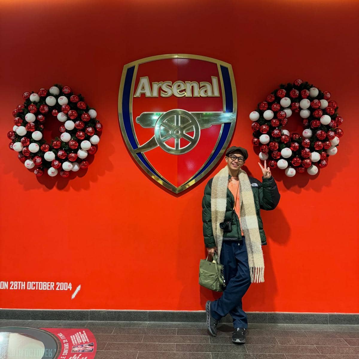 Seorang pria berpose dengan tanda kemenangan di depan logo klub sepak bola Arsenal di dinding merah dengan hiasan natal di sekitarnya.