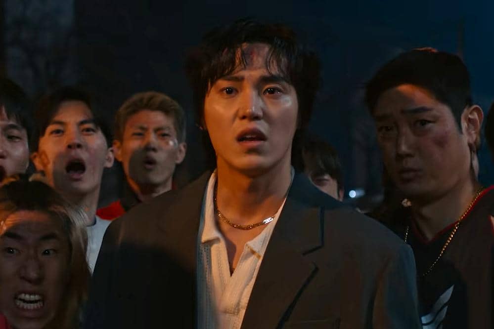 Seorang pria berdiri di depan sekelompok orang dengan ekspresi tegang pada malam hari dalam adegan drama Korea Teach You a Lesson.