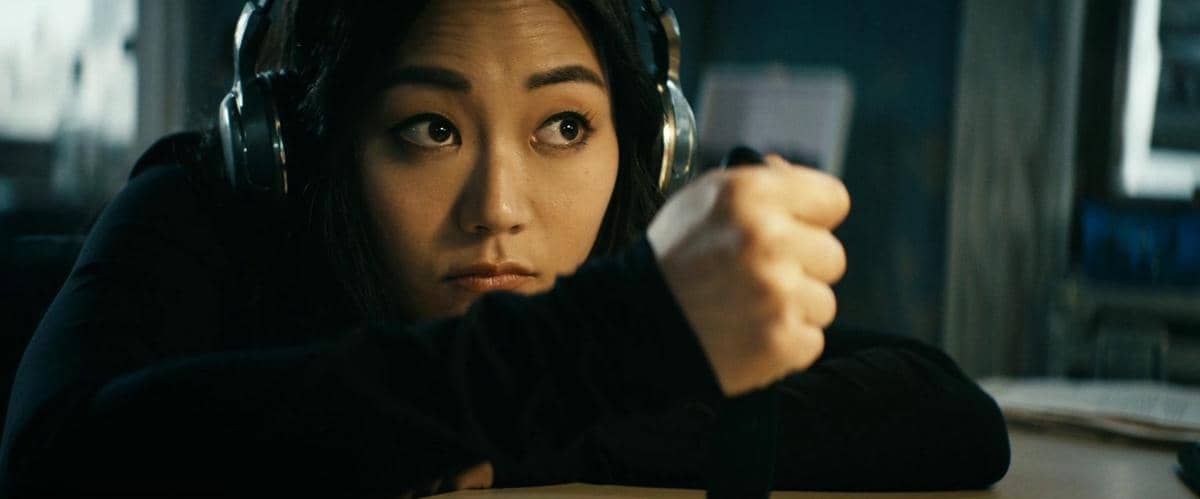 Seorang wanita mengenakan headphone besar menatap serius sambil mengangkat tangannya di dalam ruangan dengan pencahayaan redup.