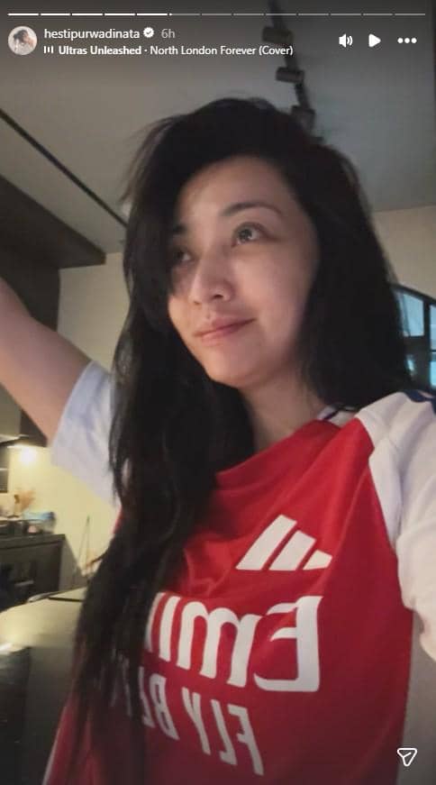 Seorang perempuan mengenakan jersey Arsenal warna merah putih tersenyum sambil mengangkat tangan di dalam ruangan.
