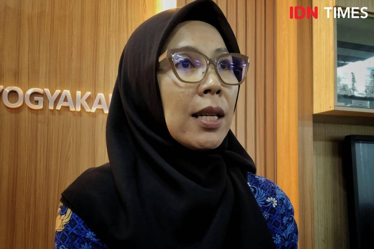 Seorang perempuan berhijab hitam dan berkacamata berbicara di ruangan dengan dinding kayu bertuliskan Yogyakarta di latar belakang.