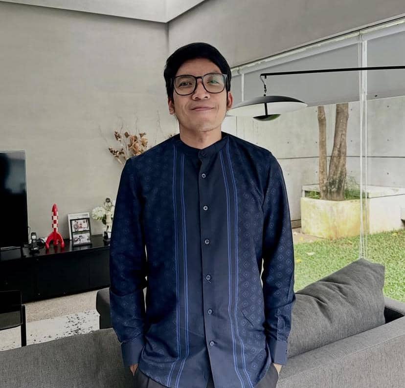 Seorang pria berkacamata mengenakan kemeja biru tua berdiri di ruang tamu modern dengan jendela besar menghadap taman.