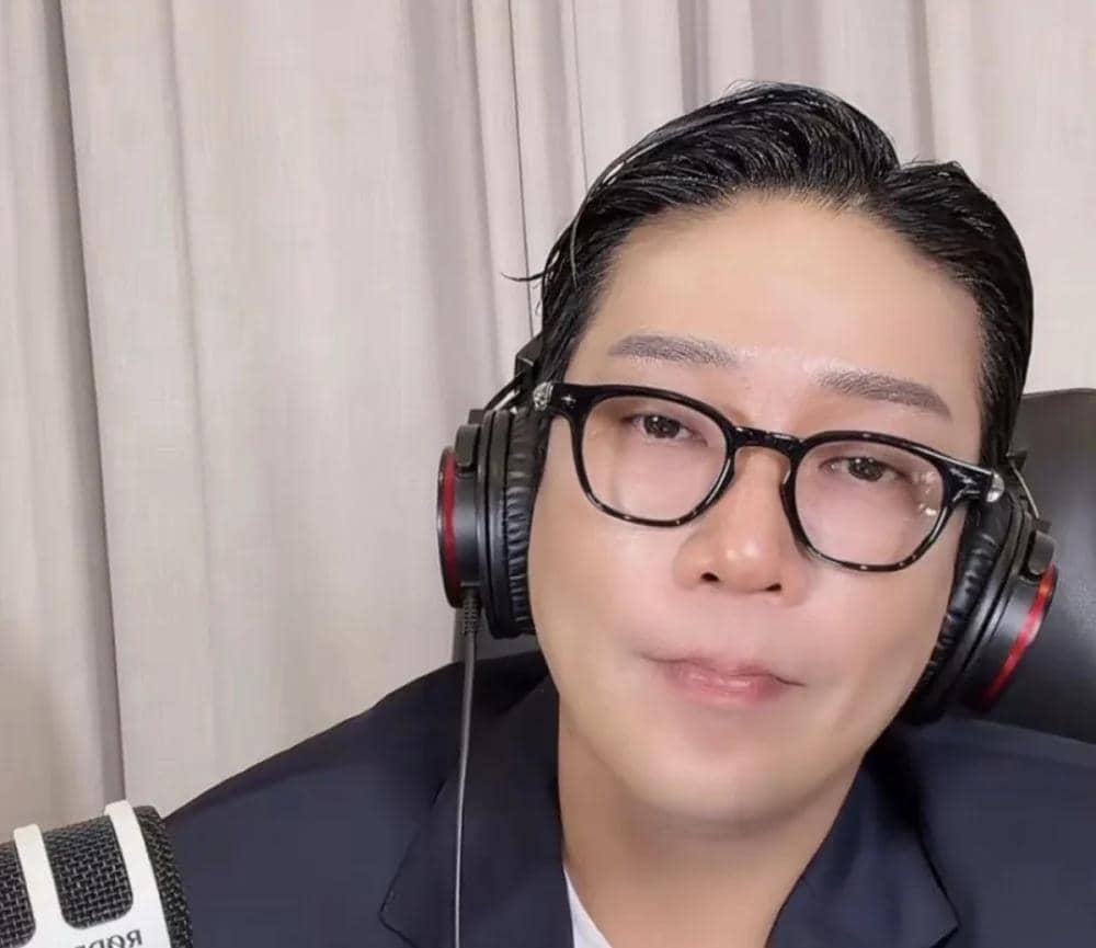 Seorang pria mengenakan kacamata hitam dan headphone besar sedang duduk di depan mikrofon dengan latar tirai berwarna terang.