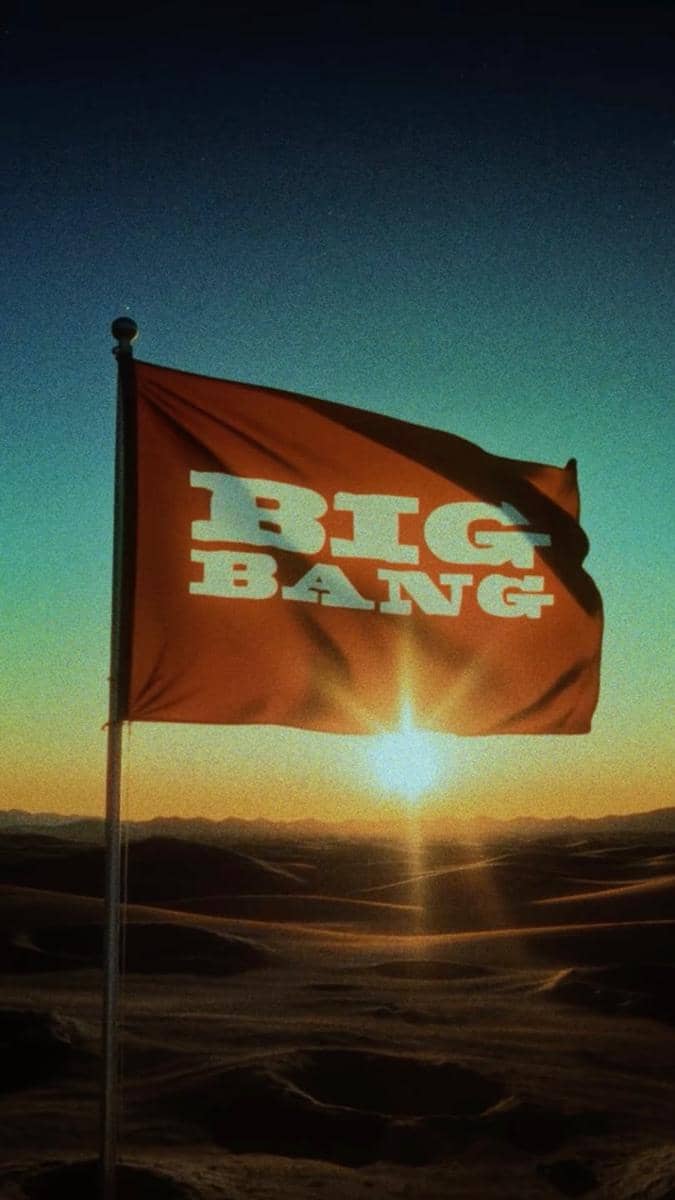 Bendera bertuliskan BIG BANG berkibar di padang pasir dengan latar matahari terbenam yang memancarkan cahaya hangat di cakrawala.